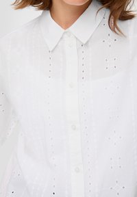Witte blouse van lichtgewicht stof met een knoopsluiting aan de voorkant, voorzien van decoratieve uitsparingspatronen en een klassiek kraagdesign.