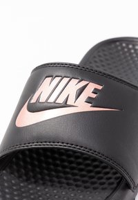 Black slides with textured footbed, featuring a wide strap adorned with a reflective rose gold Nike logo. Smooth synthetic material.