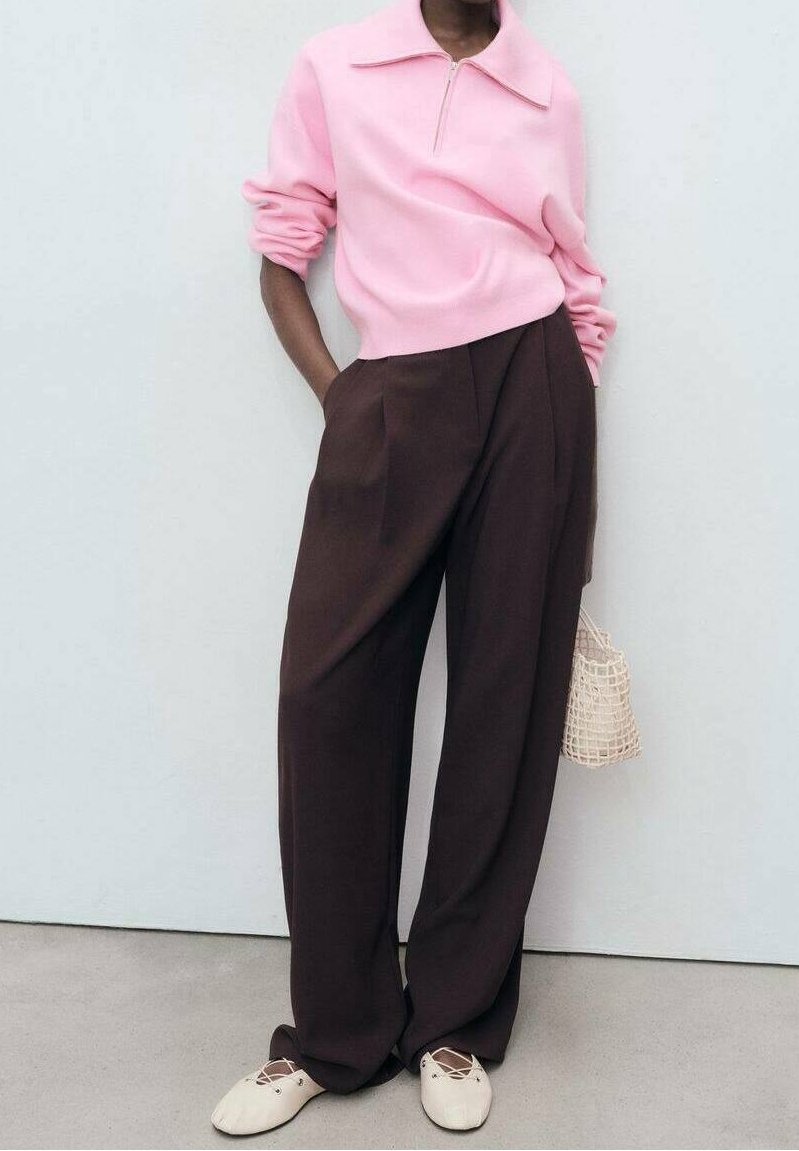 Pull-over léger rose clair avec fermeture éclair, pantalons larges taille haute marron, chaussures crème sans lacets et petit sac tissé. Tenue décontractée avec une coupe relax.