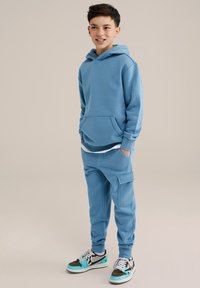 Felpa blu chiaro con tasca a marsupio, abbinata a pantaloni jogger dello stesso colore con tasca laterale. Indossata con sneakers multicolore.