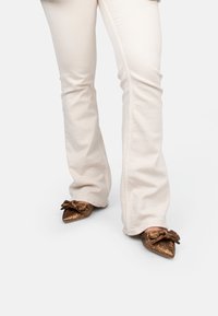 Scarpe con punta a punta in oro metallizzato, con superficie testurizzata e dettaglio a fiocco, indossate con pantaloni a palazzo chiari.