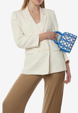 Blazer bianco in lino con due bottoni, abbinato a pantaloni beige. Regge una piccola borsa geometrica blu con accenti bianchi.