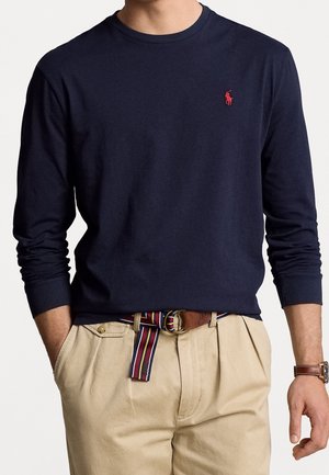 Man wearing navy long-sleeve shirt with red logo, beige pleated pants, striped fabric belt, and brown watch on left wrist.