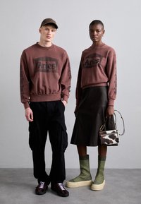 To modeller bærer matchende brune sweatshirts med "Aries" logoet. Den mandlige model har sorte cargobukser og sneakers, mens den kvindelige model har en brun lædernederdel og grønne strikkede strømper med platformsko. Hun bærer en lille, mønstret håndtaske.