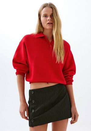 Kvinde med langt blondt hår iført en løs rød sweater og et sort asymmetrisk nederdel med nålestriber og knapper ned ad den ene side.