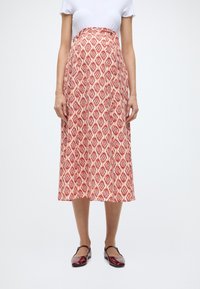 Jupe midi à motifs rouges et blancs, présentant un design en losanges, une coupe évasée et un tissu lisse. Associée à des chaussures bordeaux brillantes.