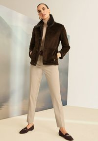 Bruine leren jas met een bontkraag, beige coltrui, beige broek en bruine loafers. Voorzien van knoopzakken en een gestroomlijnde pasvorm.