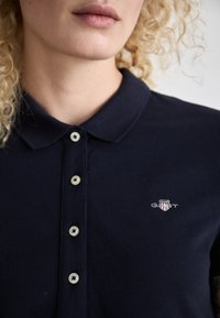 Marineblå polo-skjorte med teksturert stoff, med krage, fem knapper og en liten brodert logo på brystområdet.