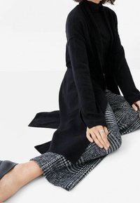 Svart lång cardigan i mjukt tyg, med avslappnad passform och öppen front. Matchas med mönstrade grå och svarta byxor, samt släta svarta skor.