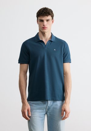 Giovane uomo che indossa una polo a maniche corte verde acqua scuro con un piccolo logo sul petto e jeans azzurri, in piedi contro uno sfondo bianco.