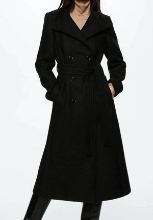 Kvinde iført en lang sort dobbeltknappet frakke med bælte, stående med hænderne i lommerne, lagdelt over en beige rullekrave.
