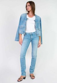 Veste en denim bleu clair avec poches avant, associée à un t-shirt blanc et un jean skinny bleu clair. Le mannequin porte des talons noirs à lanières.