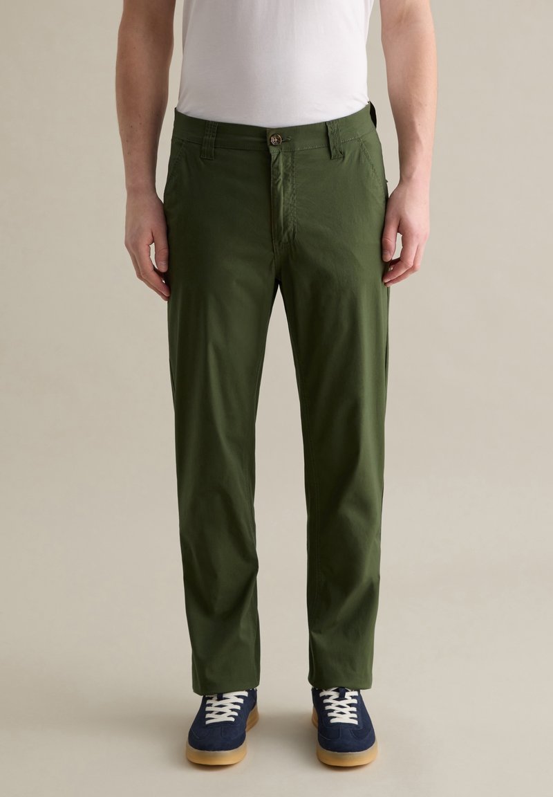 Groene katoenen broek met een rechte snit, voorzien van een knoopsluiting en zijzakken, gecombineerd met blauwe sneakers en witte veters.