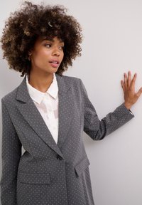 Pelēks žakets ar baltām punktītēm, strukturēts dizains, izteikti klāji un divi priekšējie kabatas, uzvilkts virs caurspīdīgas baltas blūzes ar pogu detaļām.