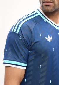 Άνδρας που φοράει μπλε αθλητική μπλούζα Adidas με ανοιχτό μπλε και λευκές ρίγες στον ώμο και στα μανίκια, και το λογότυπο της Adidas στο στήθος.