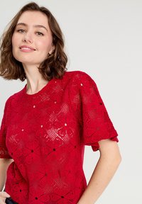 Haut en crochet rouge avec des motifs floraux, des manches courtes, un col rond et une coupe ample, mettant en valeur des détails découpés complexes.