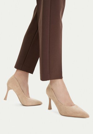 Zapatos de tacón alto beige con punta, usados con pantalones marrones a la altura del tobillo, en una persona de pie contra un fondo claro.
