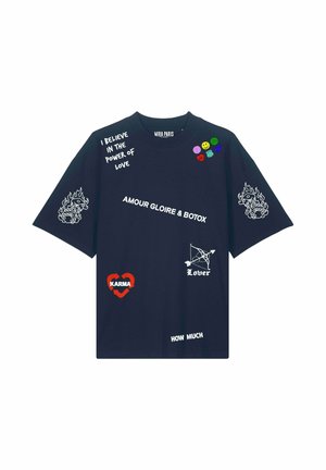 Femme aux cheveux bruns et raides portant un t-shirt oversize bleu marine avec des symboles et textes blancs et colorés, notamment "Karma," "Lover," et "Amour Gloire & Botox."