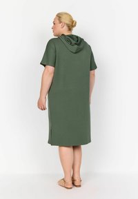 Vestido con capucha de tejido liso en color verde olivo, de longitud a la rodilla con mangas cortas y aberturas laterales para mayor comodidad de movimiento. Presenta un corte holgado y una textura suave.