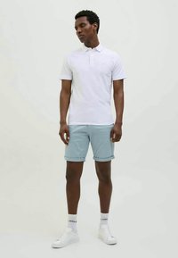 Camisa polo branca, calções azuis claros dobrados, sapatilhas brancas e meias. Modelo posa contra um fundo simples, apresentando os detalhes do conjunto de forma clara.