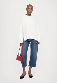 Un pull en tricot blanc à coupe décontractée, un jean droit bleu, et des escarpins noirs à bout pointu. Tient un petit sac à main rouge.