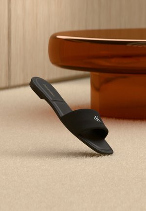 Fekete Calvin Klein papucs nyitott orral, bézs színű szőnyegen, modern beltéri környezetben, egy kerek, borostyánsárga színű asztalláb mellett pihen.