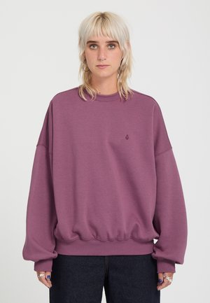 Personne aux cheveux blonds clairs portant un sweat violet large et un pantalon sombre, debout devant un fond clair uni.