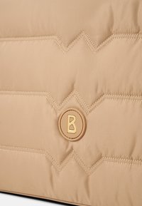 Beige puffertaske med zigzag quiltet mønster, blødt stof, guldtonet cirkulært logo med et "B" og kontraststing detail.