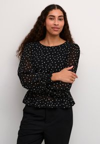 Blusa nera con pois, maniche lunghe trasparenti, polsini arricciati e orlo a peplo. Indossata con pantaloni neri, su uno sfondo semplice.