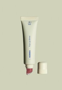 KORRES True Lip Shine dans un tube beige avec une écriture bleue et un capuchon à vis, présentant une texture douce et soyeuse rosa à l'ouverture.