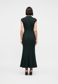 Γυναίκα που στέκεται με την πλάτη στην κάμερα φορώντας εφαρμοστό, αμάνικο, μεσαίο φόρεμα σε σκούρο πράσινο χρώμα και μαύρες ψηλοτάκουνες πέδιλα με λουράκια σε απλό φόντο.