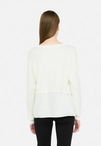 Top bianco a maniche lunghe con design crop, caratterizzato da una texture morbida e liscia e un effetto a strati sul retro. Abbinato a pantaloni neri.