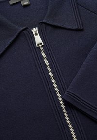 Marineblauwe ritsjack met een gestructureerde stof, puntige kraag en zilveren ritsdetails. Kenmerkt zich door strakke lijnen en schone naden.