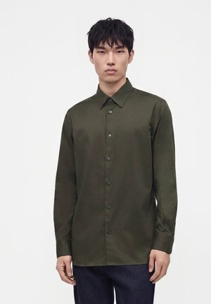Jeune homme aux cheveux noirs courts portant une chemise verte olive à manches longues boutonnée et un pantalon sombre sur un fond blanc uni.