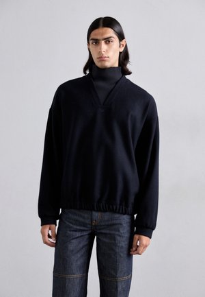 Zwarte oversized trui met een V-hals en ribbelige col, met een gathered zoom en lange mouwen, gedragen over donkere spijkerbroek.