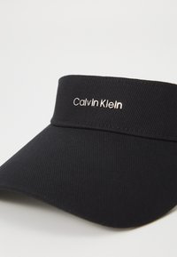 Zwarte pet met een gestructureerde stof, met een metalen "Calvin Klein" logo op de voorkant. De rand is breed en gebogen.