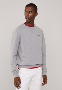 Felpa grigia con polsini e orlo a coste, caratterizzata da un piccolo logo ricamato. Indossata sopra una t-shirt rossa a girocollo. Pantaloni bianchi.