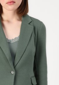 Olivově zelený blazer se strukturovanými klopami, zapínáním na jeden knoflík a texturovaným materiálem. Pod ním šedý top s krajkovým lemem viditelný.