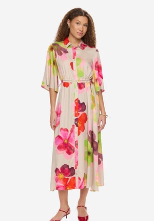 Kvinde iført en beige midikjole med store farverige blomsterprint, halvlange ærmer, knaplukning foran, bindebånd i taljen og røde hælede sandaler.