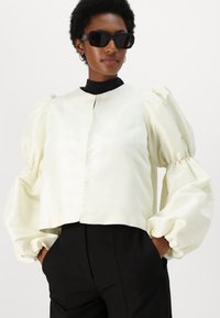 Blouse cropped en satin crème avec des manches bouffantes volumineuses et un col rond, associée à un pantalon noir. Le modèle porte des lunettes de soleil.