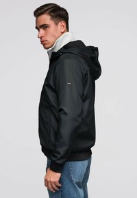 Młody mężczyzna w czarnej kurtce z kapturem i niebieskich dżinsach, patrzący w lewo na jednolity, jasnoszarym tle.