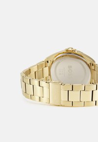 Montre-bracelet en métal doré avec un bracelet à maillons, un boîtier arrondi, et un dos transparent mettant en valeur le mécanisme de la montre. Nom de la marque gravé.