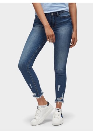 Pantalones vaqueros ajustados en denim azul medio, con detalles rasgados en el dobladillo, combinados con zapatillas blancas. Textura suave y diseño ceñido.