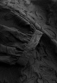 Tessuto nero con motivo a foglie in rilievo, caratterizzato da sezioni trasparenti e volant strutturati lungo i bordi. Finitura liscia e dettagliata.