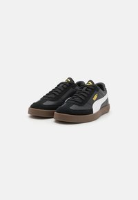 Svart sneakers laget av semsket og lær, med en hvit stripe og gule logoaksenter, samt en brun gummisåle.