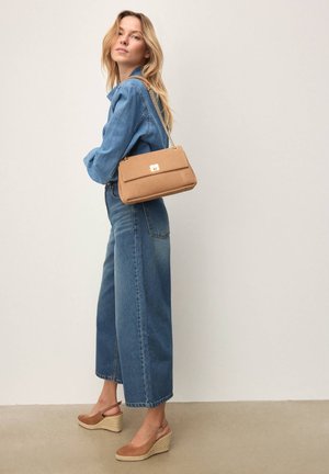 Beżowa torebka na ramię z złotym zapięciem, w kształcie prostokąta, z łańcuszkowym paskiem. Noszona z dżinsową koszulą i szerokimi dżinsami; całość uzupełniają buty na koturnie.