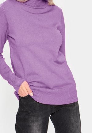 Personne portant un pull à col roulé violet et un jean noir, avec une main dans la poche avant du jean.