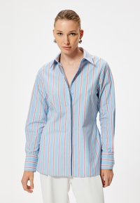 Camisa abotonada de color azul claro con rayas rojas verticales, de mangas largas y con un collar clásico. Textura de tela suave con un corte ajustado.
