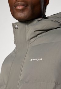 Gray puffer jacket with a high collar, zippers, and branded embroidery. Smooth texture with stitched detailing.