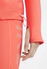 Top atlético de color coral brillante con mangas texturizadas y dobladillo ajustable, combinado con leggings ceñidos de color coral que presentan rayas verticales blancas.